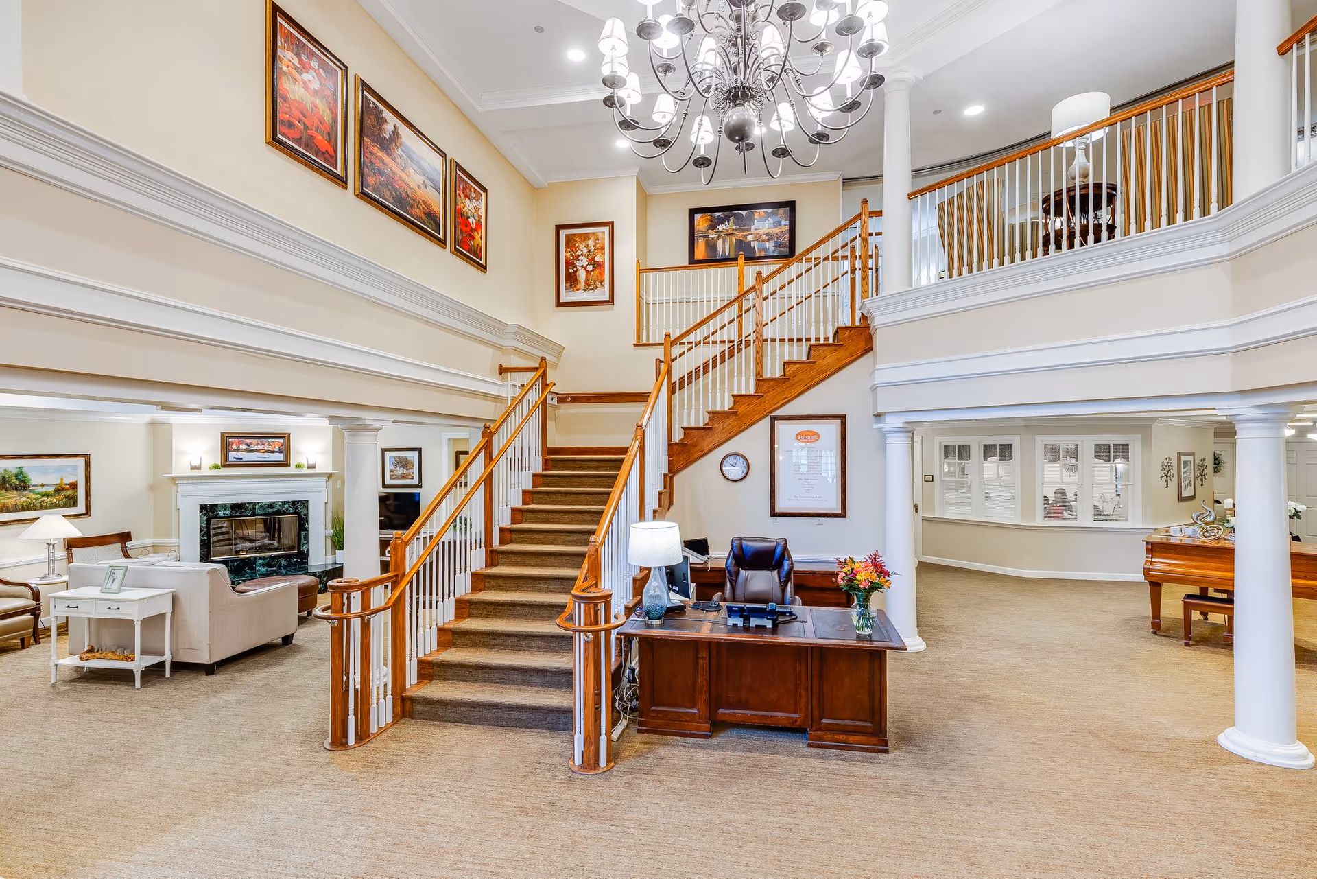 Interior view of a senior living facility lobby with a wooden staircase in the center, a dark wood reception desk with a lamp and flowers, beige carpet, white columns, a chandelier, and several framed paintings on the walls. There is a seating area with sofas and a fireplace to the left and a piano to the right.