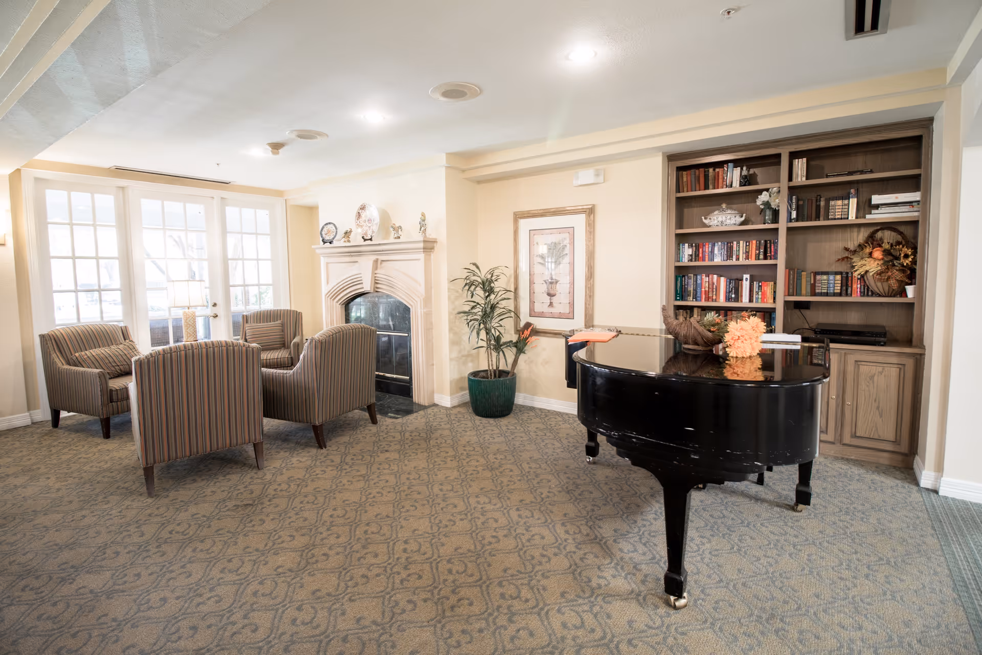 A cozy common area with a black grand piano adorned with a floral arrangement, a built-in wooden bookshelf filled with books and decorative items, a framed artwork on the wall, a potted plant, a fireplace with decorative plates and figurines on the mantel, and four striped armchairs arranged around a small table near glass double doors letting in natural light.