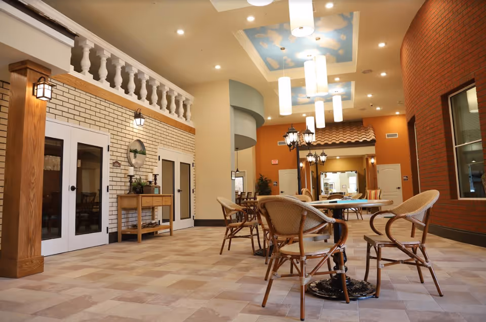 Interior view of a senior living facility common area with wicker chairs and round tables on a tiled floor. The walls feature a mix of white brick, orange paint, and red brick. The ceiling has decorative light fixtures and a painted sky with clouds. There is a small wooden console table against the wall and a streetlamp-style light fixture near the tables.