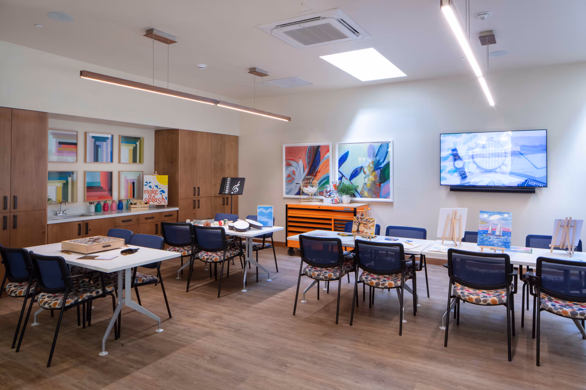 A bright and spacious activity room with two white tables surrounded by chairs with patterned cushions. The tables have art supplies, small paintings on easels, and musical instruments. The room features wooden cabinets, colorful abstract artwork on the walls, a large flat-screen TV, and a skylight providing natural light.