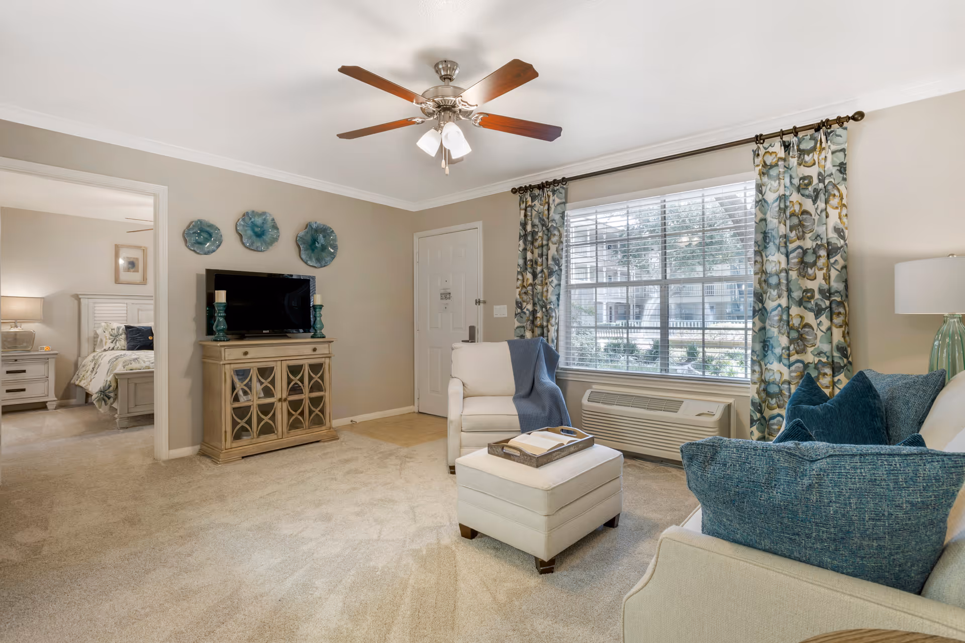 A cozy living room with beige walls and carpet, featuring a white armchair with a blue throw, a matching ottoman with a tray and book, a sofa with blue cushions, a wooden TV stand with a flat-screen TV and decorative teal plates on the wall above. A large window with floral curtains lets in natural light, and a ceiling fan with lights is mounted on the ceiling. The room opens into a bedroom with a white bed and nightstand visible.