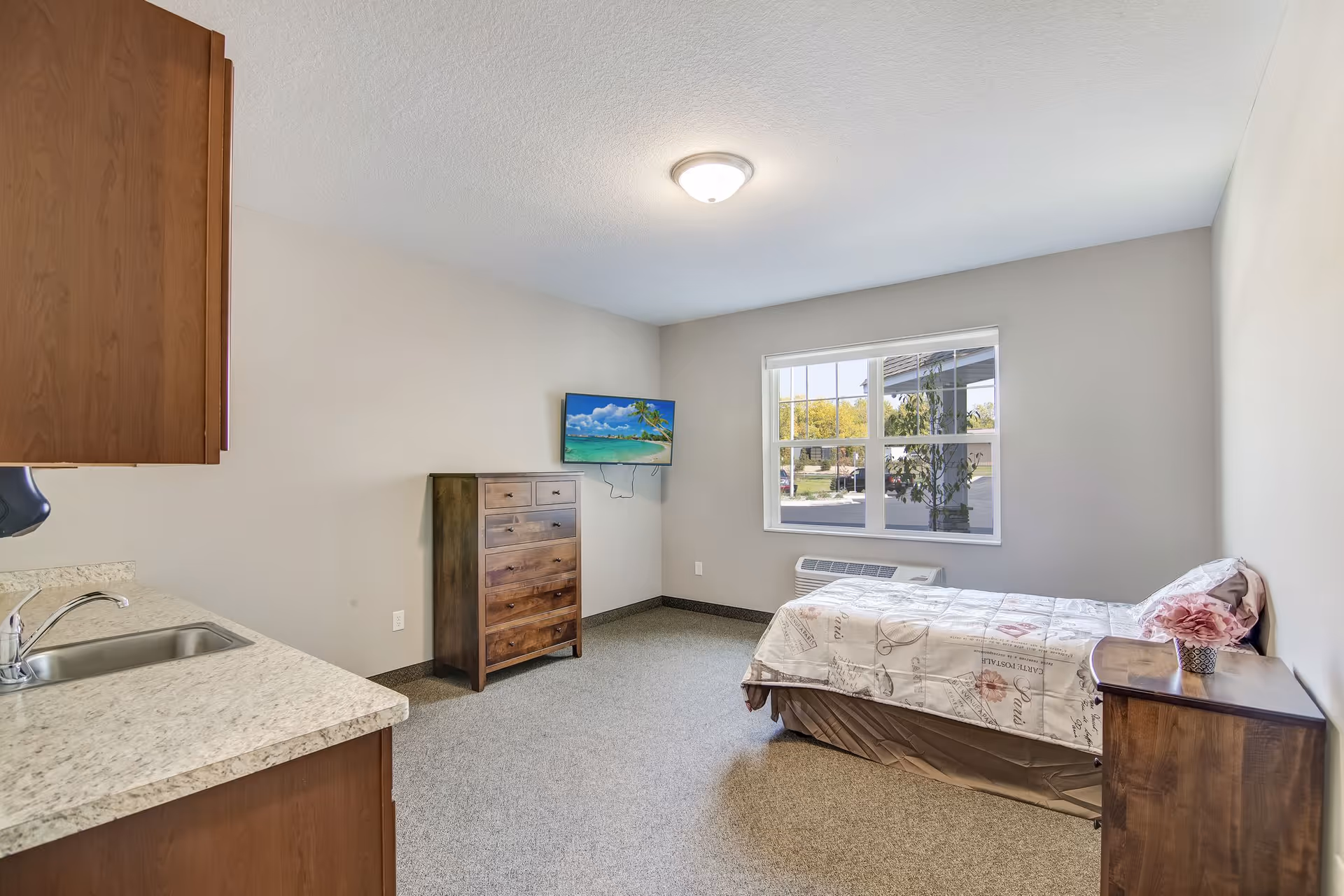 A simple, clean bedroom in an assisted living facility featuring a single bed with floral bedding, a wooden nightstand with a flower vase, a wooden dresser, a wall-mounted flat-screen TV, a window with a view of the outside, and a small kitchenette area with a sink and countertop.