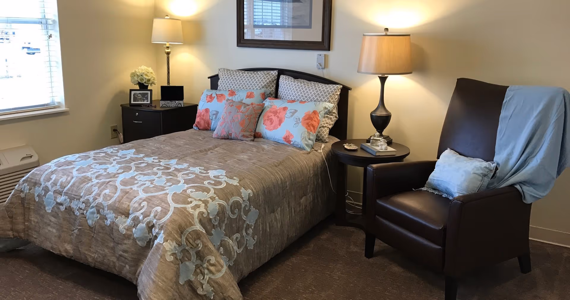 Neatly made bedroom with a bed dressed in patterned bedding, two bedside tables with lamps, and a leather armchair with a throw.