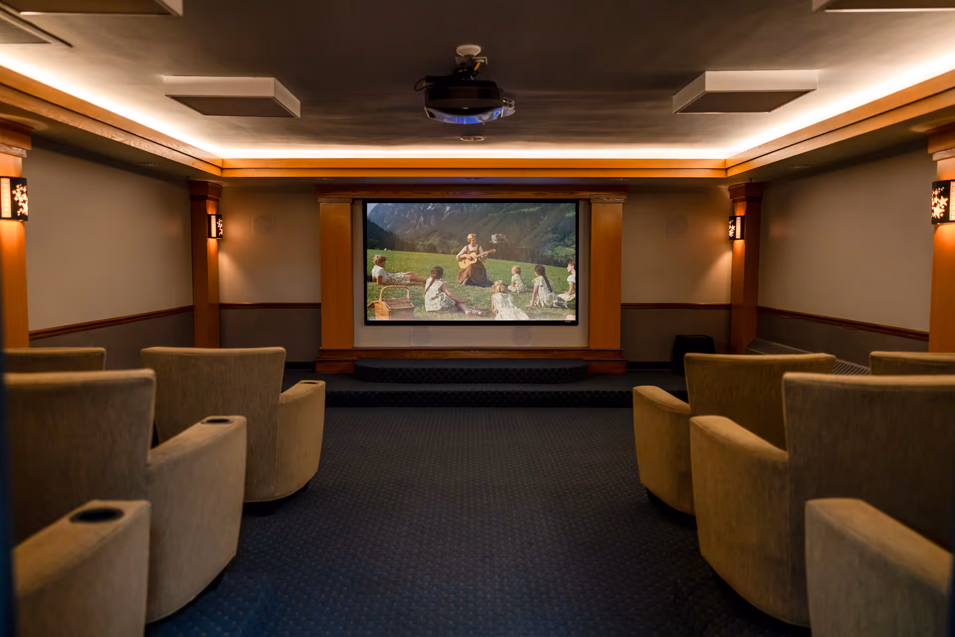 A cozy home theater room with beige upholstered recliner chairs arranged in rows facing a large screen showing a scene from a movie. The room has soft lighting with wall sconces and a ceiling-mounted projector.