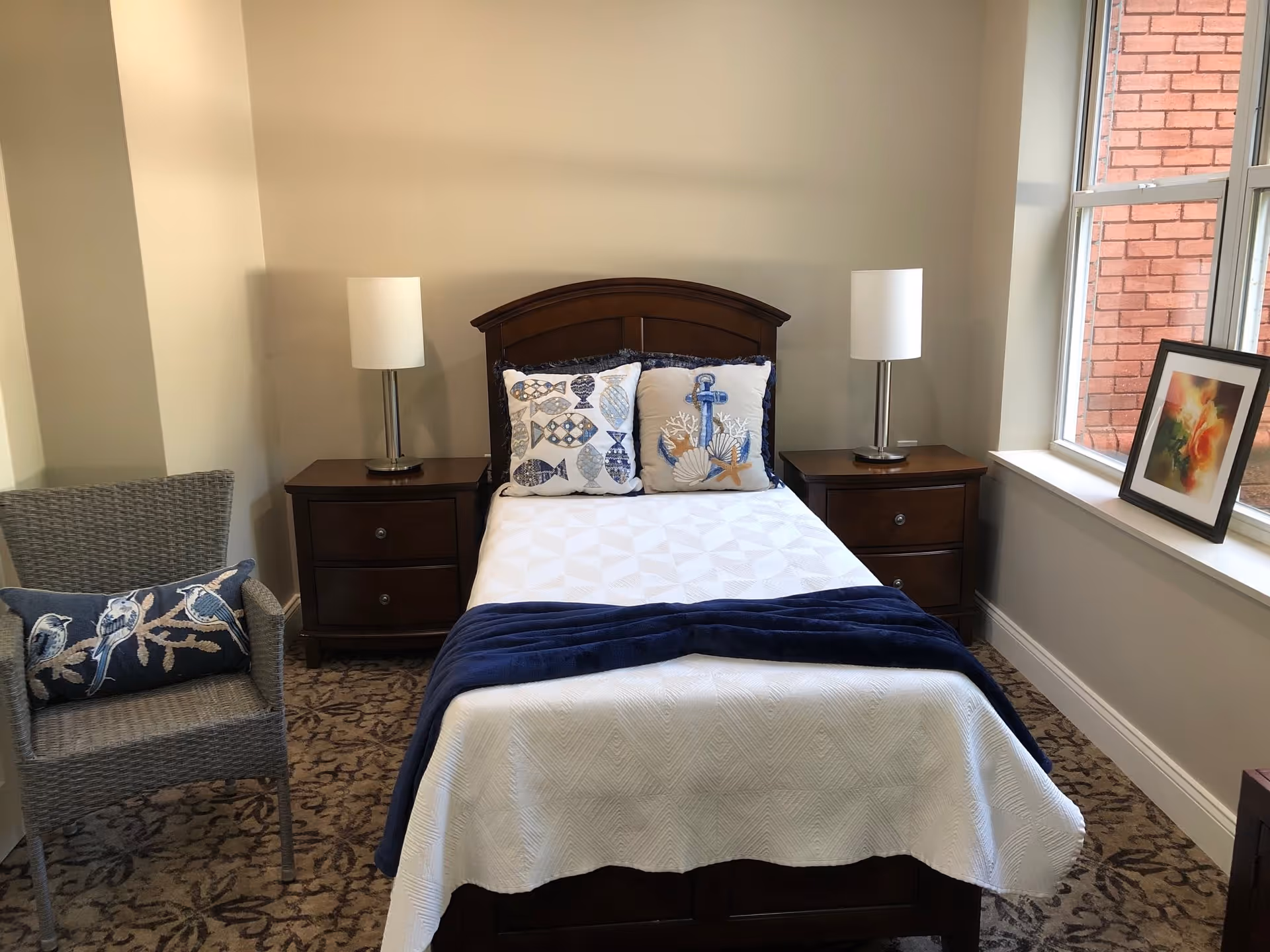 A neatly made single bed with a white quilt and a dark blue throw blanket at the foot. The bed has two decorative pillows with fish and nautical designs. On either side of the bed are dark wooden nightstands with modern white lamps. To the left is a wicker chair with a blue cushion featuring bird designs. To the right is a window with a framed floral picture on the windowsill. The room has beige walls and patterned carpet flooring.