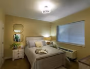 A cozy bedroom with a neatly made bed featuring multiple pillows and a tray on top. There is a nightstand with a lamp and a plant beside the bed. Two decorative mirrors hang on the wall above the nightstand. A window with closed blinds is on the right wall, and an air conditioning unit is below the window. The room has soft yellow walls and carpeted flooring.