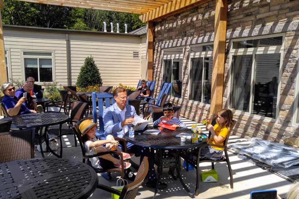 An outdoor patio area at The Pearl at Watkins Centre - Memory Care Community with several people sitting at round tables under a wooden pergola. The area is furnished with black metal tables and chairs. Some people are engaged in conversation, while others are reading or using their phones. The patio is adjacent to a building with stone and siding walls and multiple windows.