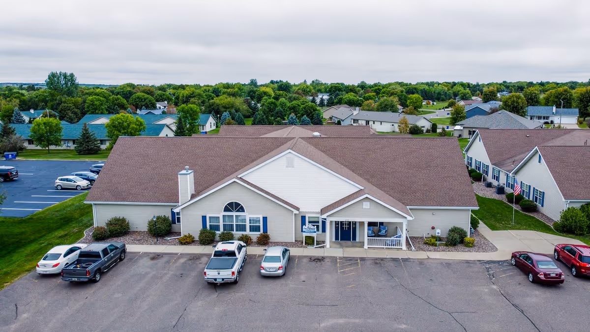 Aerial view of a single-story senior living facility building with a brown roof and beige siding, surrounded by a parking lot with several cars parked. The building has a small covered entrance with a porch and blue shutters on the windows. The surrounding area includes green lawns, trees, and neighboring residential houses under a cloudy sky.
