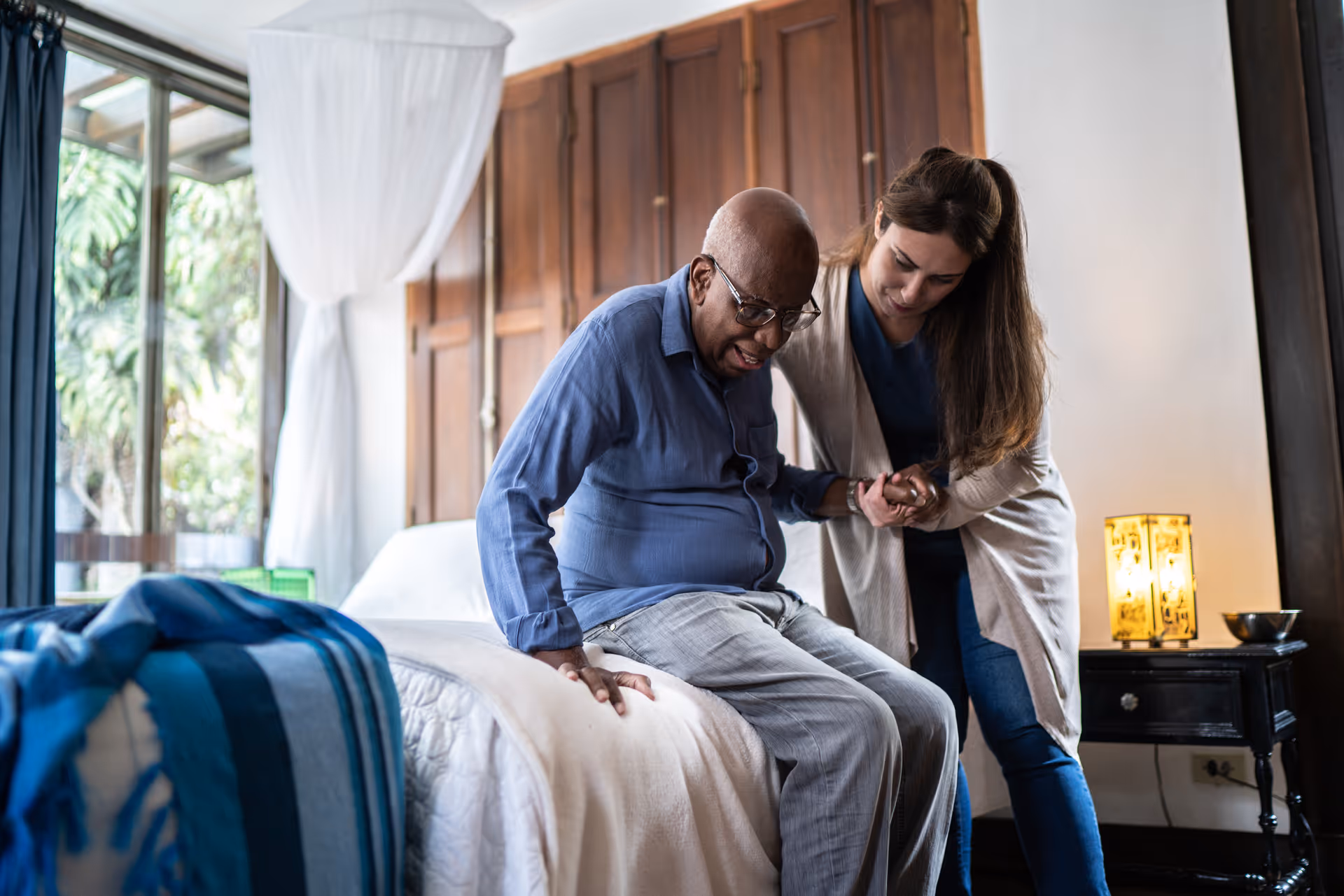 A caregiver assists an elderly man as he sits on the edge of a bed in a well-lit bedroom with wooden furniture and a window with white curtains.