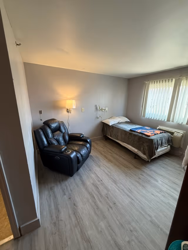 Small room with a single bed, a black recliner, a wall lamp, and a window with vertical blinds.