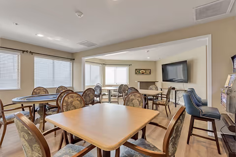 Communal dining/activity room with multiple tables and upholstered chairs, large windows, and a wall-mounted TV.