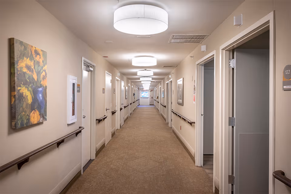 A long, well-lit hallway in a senior living facility with beige walls and carpeted floor. The hallway has multiple doors on both sides, handrails along the walls, ceiling lights, and framed artwork hanging on the walls.