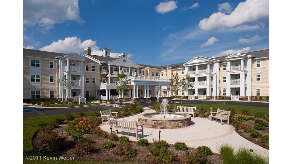 Three-story senior living building with a landscaped courtyard, fountain and benches under a blue sky.