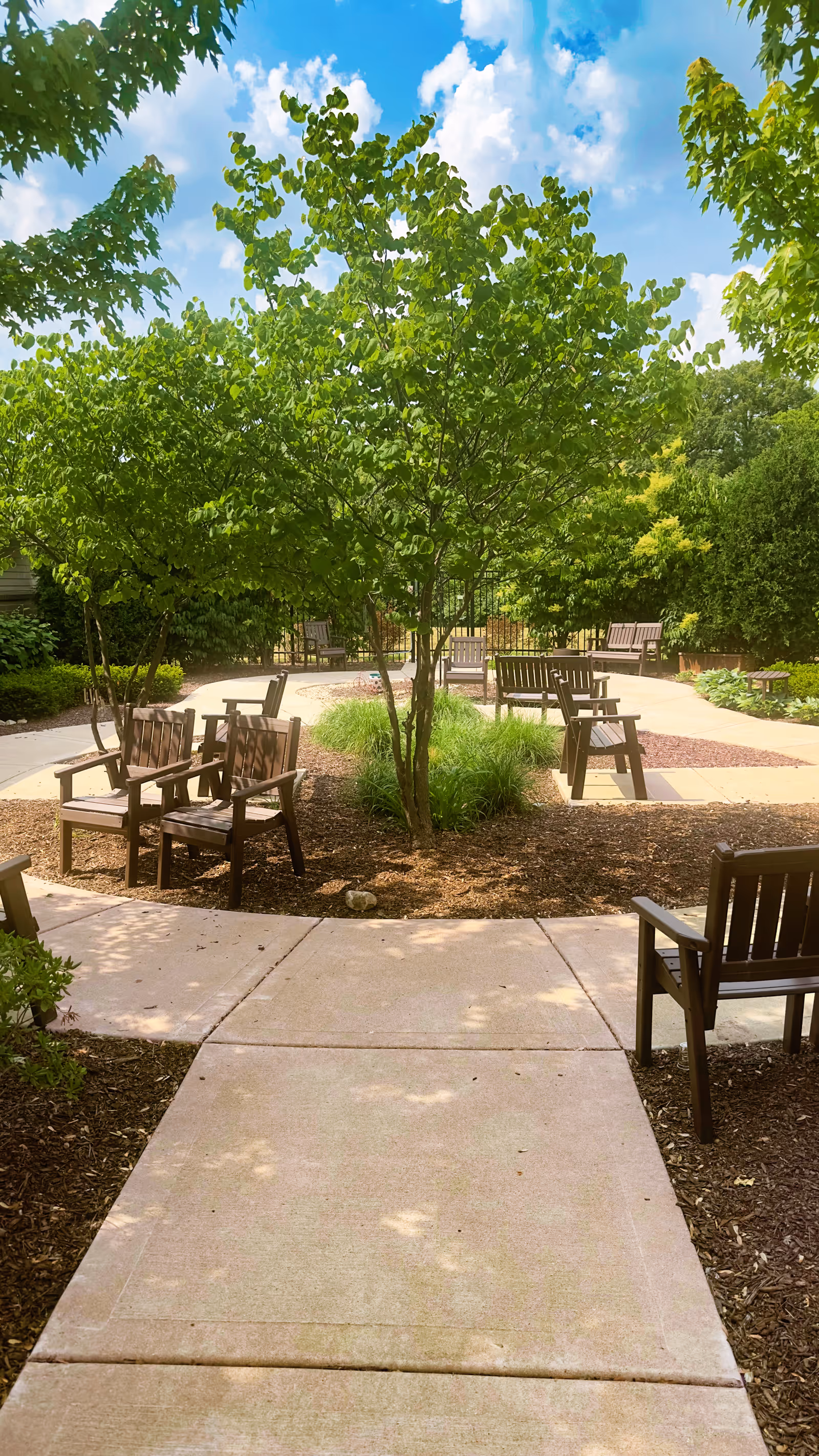 Outdoor garden area with multiple wooden chairs arranged around a circular paved pathway, surrounded by green trees and bushes under a partly cloudy blue sky.