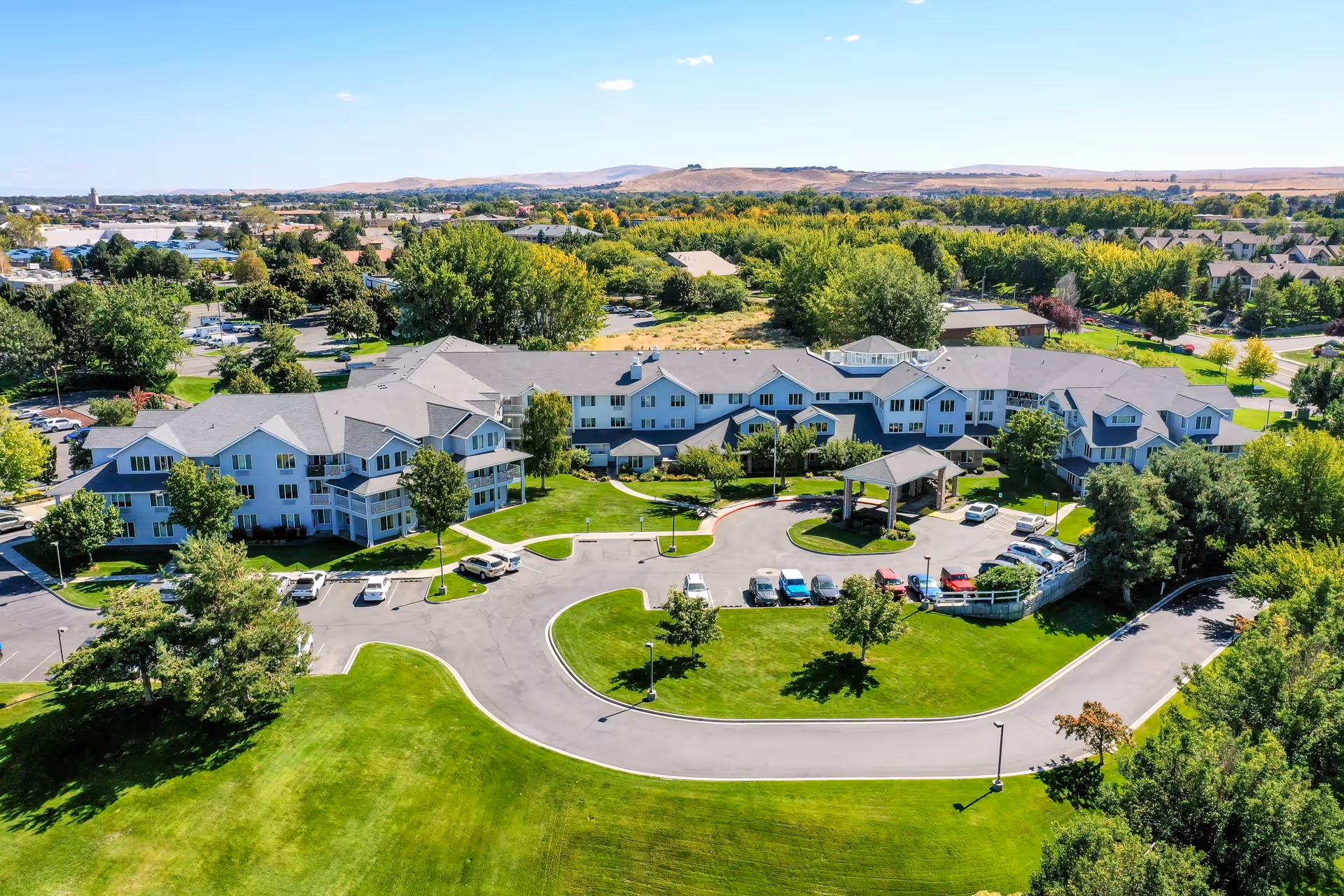 Aerial view of Solstice Senior Living at Kennewick, showing a large, multi-wing building surrounded by green lawns, trees, and a circular driveway with parked cars. The background includes more trees, residential areas, and distant hills under a clear blue sky.