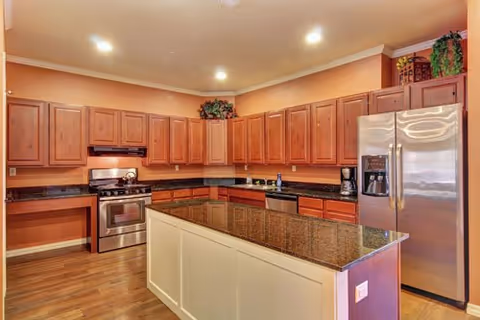 A spacious kitchen with wooden cabinets, a large granite countertop island in the center, stainless steel appliances including a refrigerator and stove, and recessed ceiling lights. There are decorative plants placed above the cabinets.