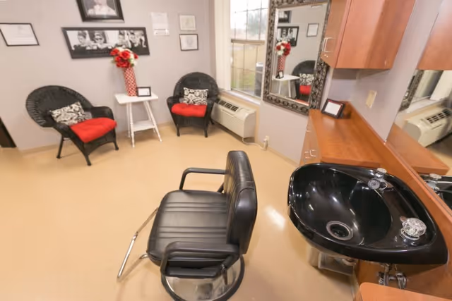 A salon-style interior with a black barber chair and wash sink, two wicker chairs with red cushions, a mirror and cabinetry.
