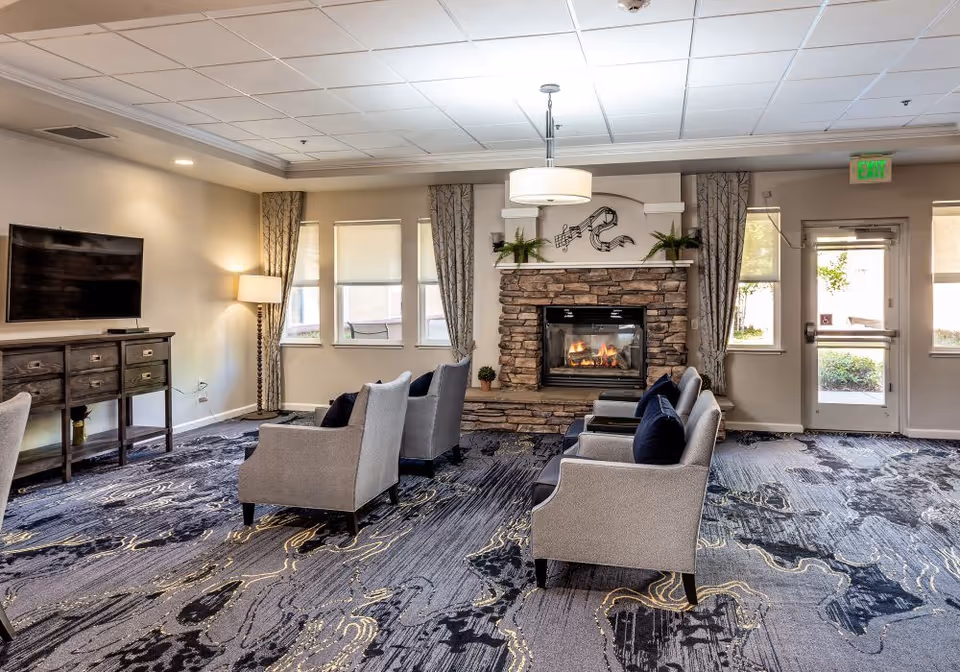 A cozy senior living facility lounge area with a stone fireplace lit with a fire. There are four gray armchairs with dark blue cushions arranged facing the fireplace. The room has a patterned carpet in shades of gray and blue, a wooden console table with a flat-screen TV mounted above it, and a floor lamp in the corner. Windows with curtains and a glass door letting in natural light are visible in the background.
