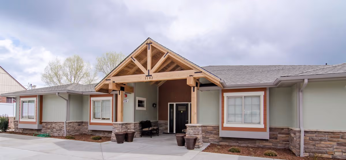 Front exterior of a single-story assisted living building with a covered wooden gable entry, stone veneer, and surrounding pavement.