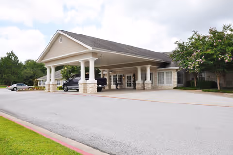 Front entrance of a senior living building with a covered porte-cochere, vehicles parked beneath it, and landscaped grounds.