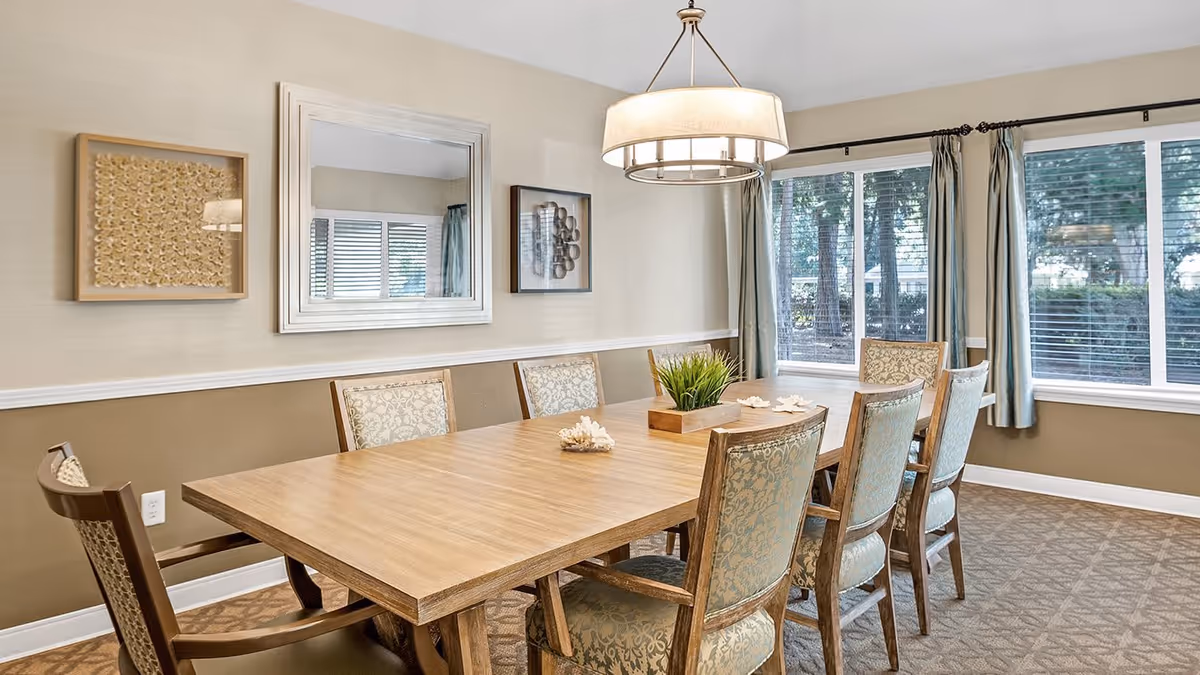 A dining room with a long wooden table surrounded by eight upholstered chairs with patterned fabric. The walls are painted beige with white trim, and there are three framed decorative pieces hanging on the wall, including a large mirror in the center. A modern chandelier hangs above the table. Large windows with curtains allow natural light to fill the room, showing trees outside.