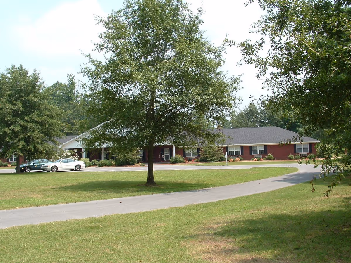 A single-story brick building with a dark roof, surrounded by green grass and trees. There is a curved driveway in front of the building with two parked cars. The scene is outdoors with a clear sky.