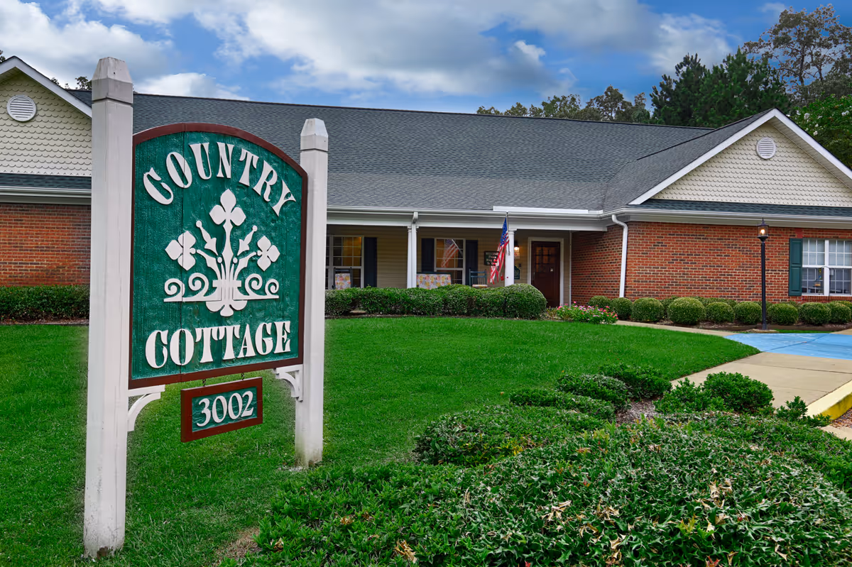 Exterior view of a single-story brick building with a green lawn and bushes in front. A large wooden sign in the foreground reads 'Country Cottage 3002'. The building has a gray shingled roof, white trim, and an American flag near the entrance.