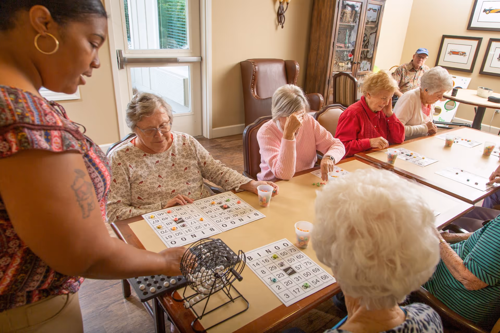 A group of elderly people sitting around a table playing bingo with a staff member assisting by operating the bingo cage. The room is well-lit with natural light from a window and has comfortable chairs and framed pictures on the wall.