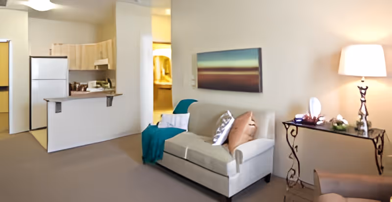 Open-plan living area with a small sofa and throw, side table with lamp, and a kitchenette with refrigerator in the background.