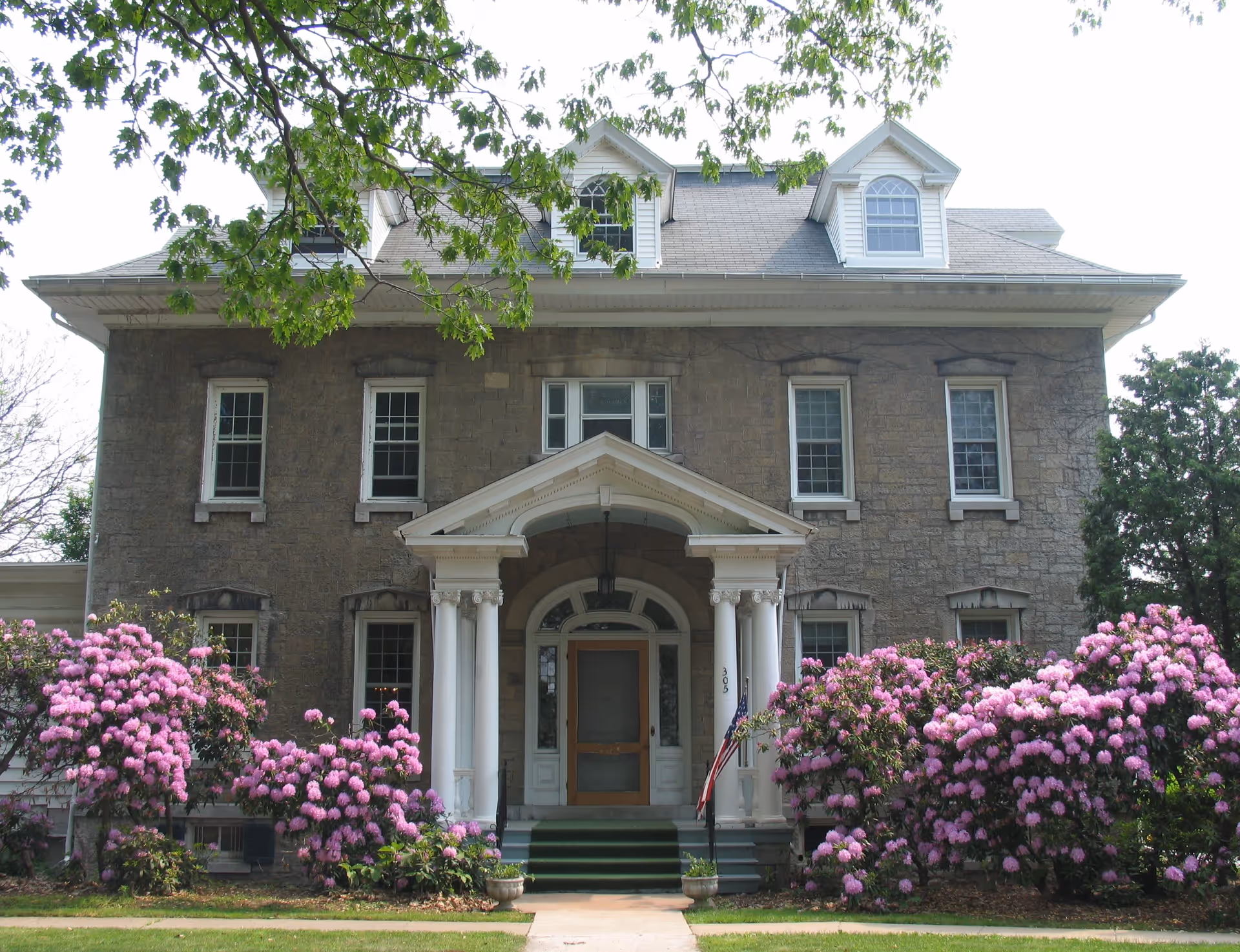 Stone two-story building with a central columned portico entrance, dormer windows on the roof, and large pink flowering shrubs at the front.