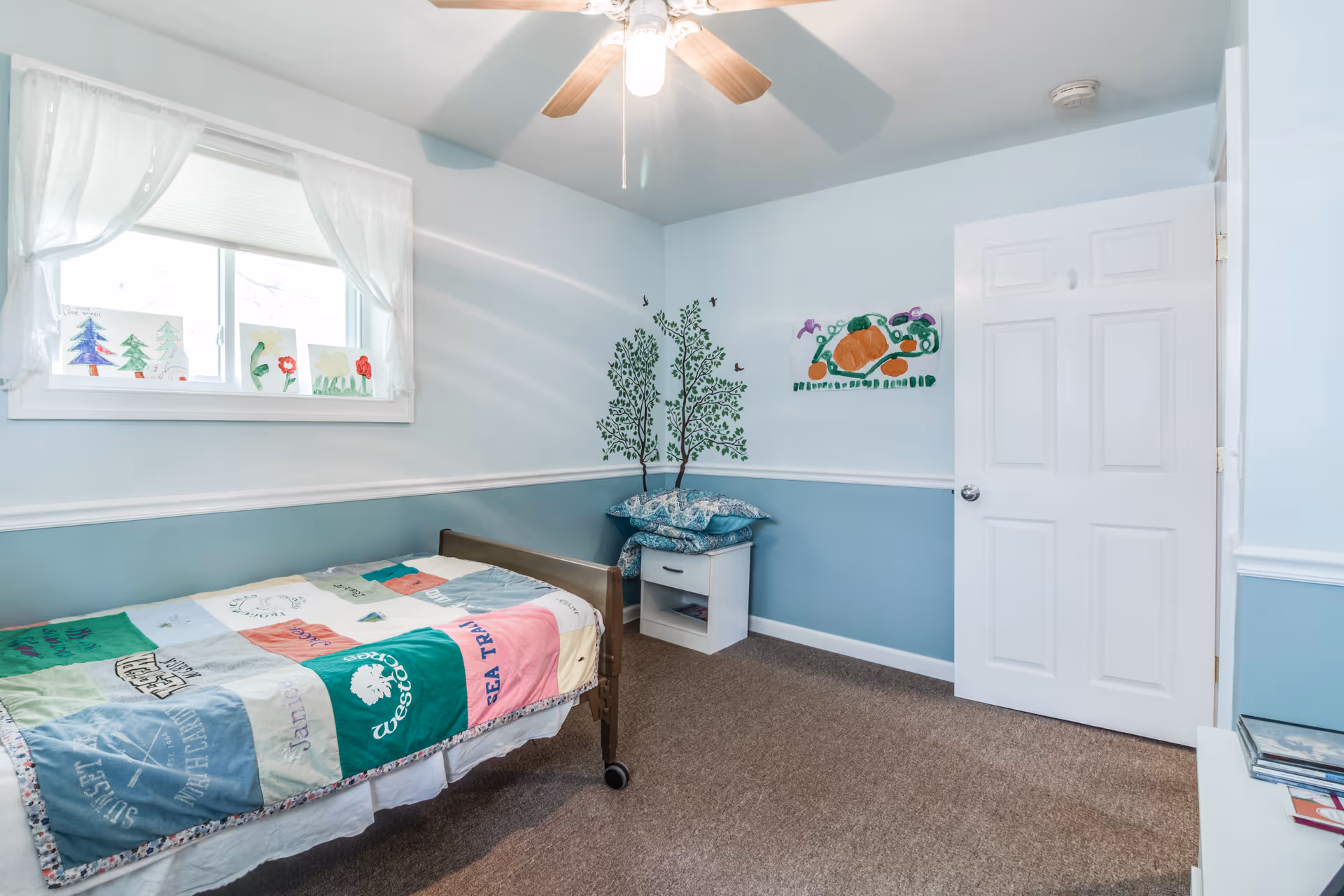 A small bedroom with light blue walls and a white ceiling fan. There is a single bed with a colorful patchwork quilt, a white nightstand with pillows stacked on top, and children's artwork displayed on the wall and windowsill. The room has a beige carpet and a white door that is partially open.