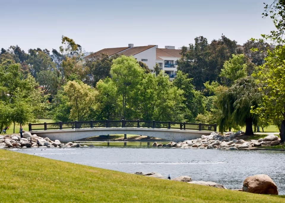 A peaceful outdoor scene featuring a small arched bridge over a pond surrounded by green trees and grass, with a building partially visible in the background.