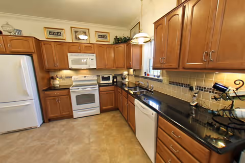 A spacious kitchen with wooden cabinets, a white refrigerator, a white oven with a microwave above it, a dishwasher, and a black countertop. The kitchen has beige tiled flooring and a window above the sink letting in natural light. There are various kitchen appliances and decorative items on the counters and cabinets.