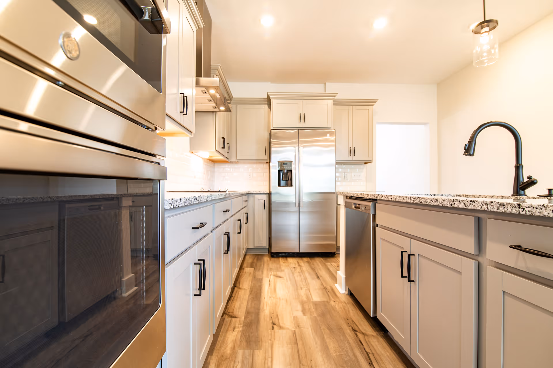 Bright modern kitchen featuring stainless steel appliances, light cabinets, a central island with a black faucet, and wood-look flooring.