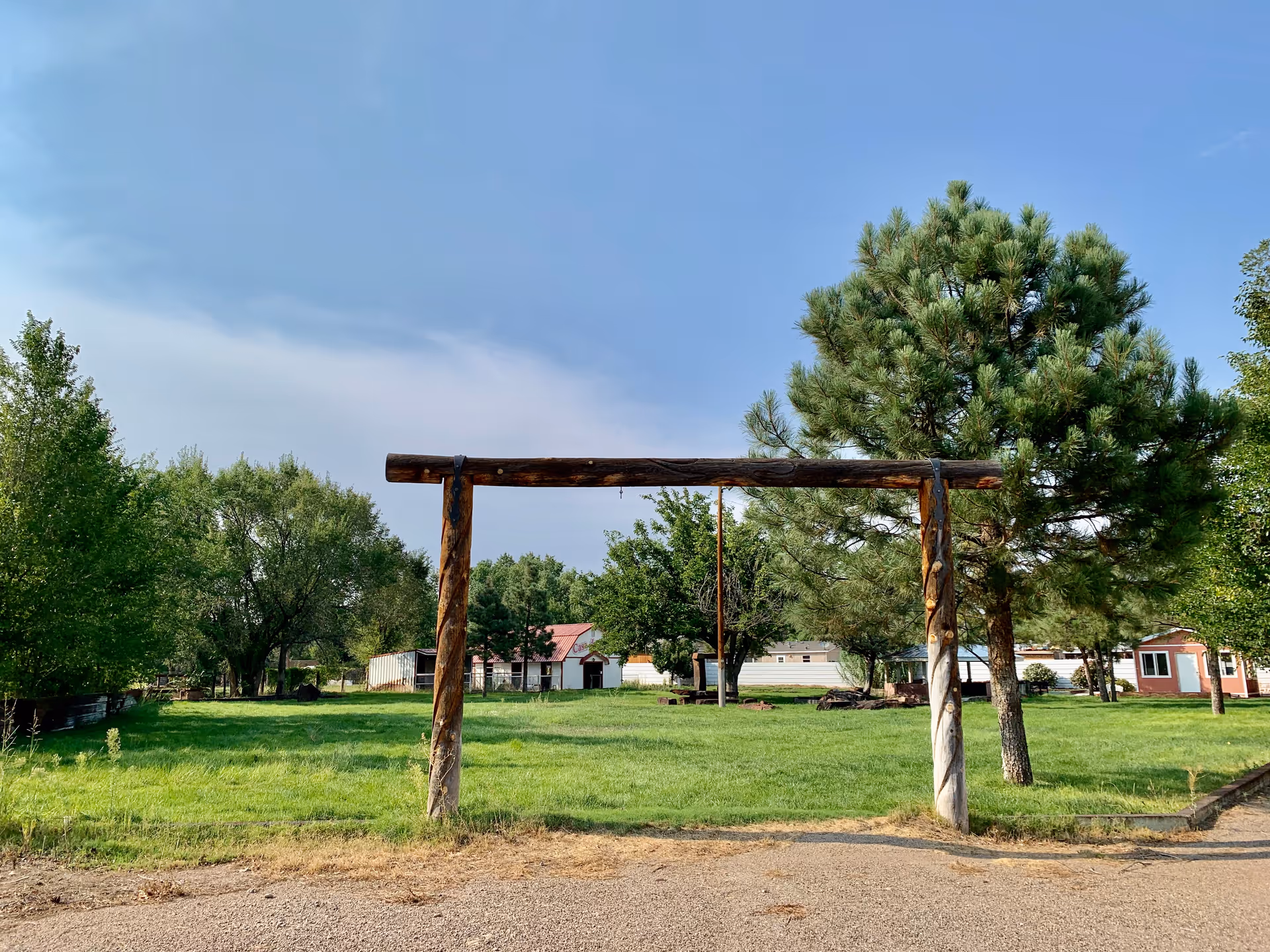 A rustic wooden gateway stands over a grassy yard with trees and small buildings under a clear blue sky.