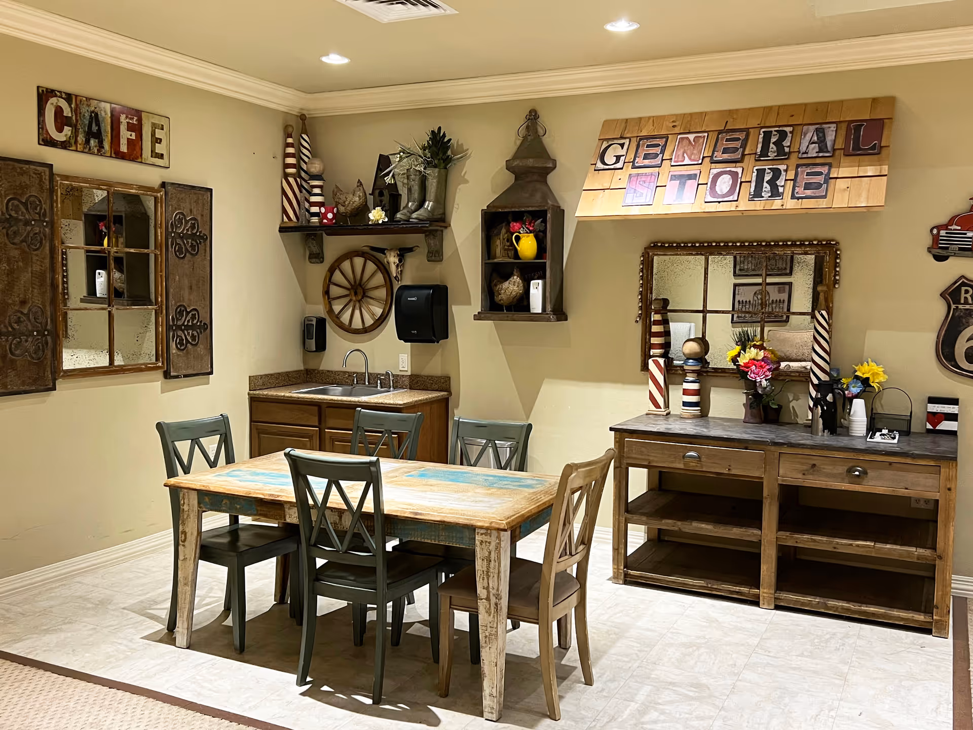 A cozy dining area with a rustic wooden table and four chairs. The walls are decorated with vintage-style signs reading 'CAFE' and 'GENERAL STORE'. There is a small sink with a cabinet underneath, shelves with decorative items including a wagon wheel, and a wooden sideboard with flowers and other decor. The floor is tiled and the lighting is warm.