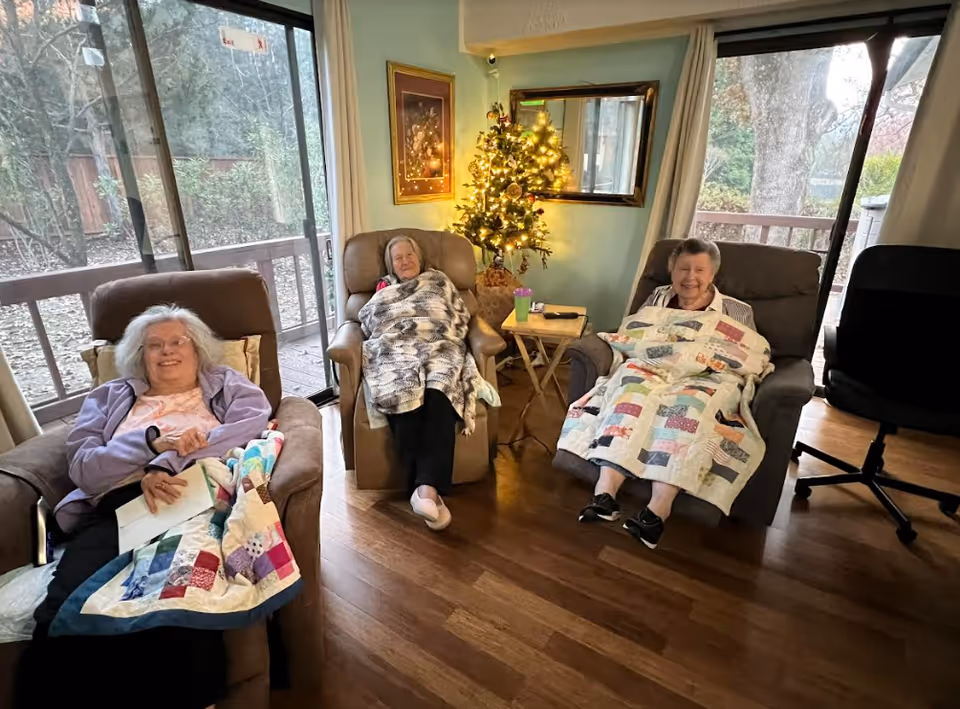 Three elderly women sitting comfortably in recliner chairs inside a cozy living room. Each woman is covered with a colorful quilt. Behind them is a small decorated Christmas tree with lights, a framed painting on the wall, and a large window showing an outdoor view with trees and a wooden deck.