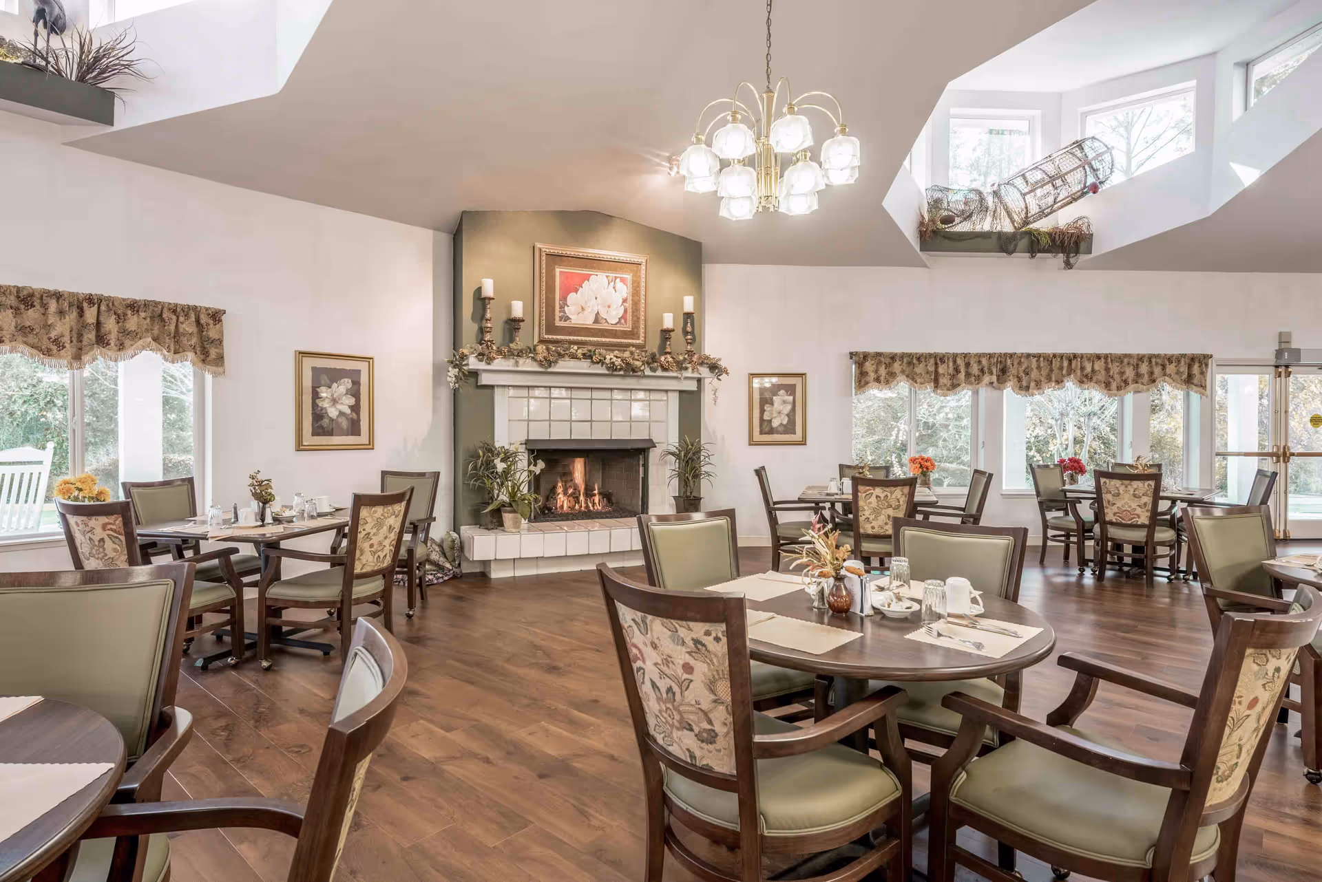 A cozy dining room with multiple round tables and chairs arranged neatly. The room features a lit fireplace with decorative candles and framed floral artwork above it. Large windows with floral valances allow natural light to fill the space, and a chandelier hangs from the ceiling. The floor is wooden, and there are plants and decorative items placed around the room.