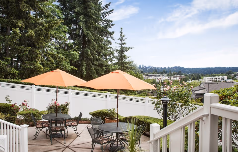Outdoor patio area with two round metal tables, each shaded by an orange umbrella. The patio is surrounded by green bushes, flowering plants, and a white privacy fence. Tall evergreen trees and a distant view of buildings and hills are visible under a partly cloudy sky.