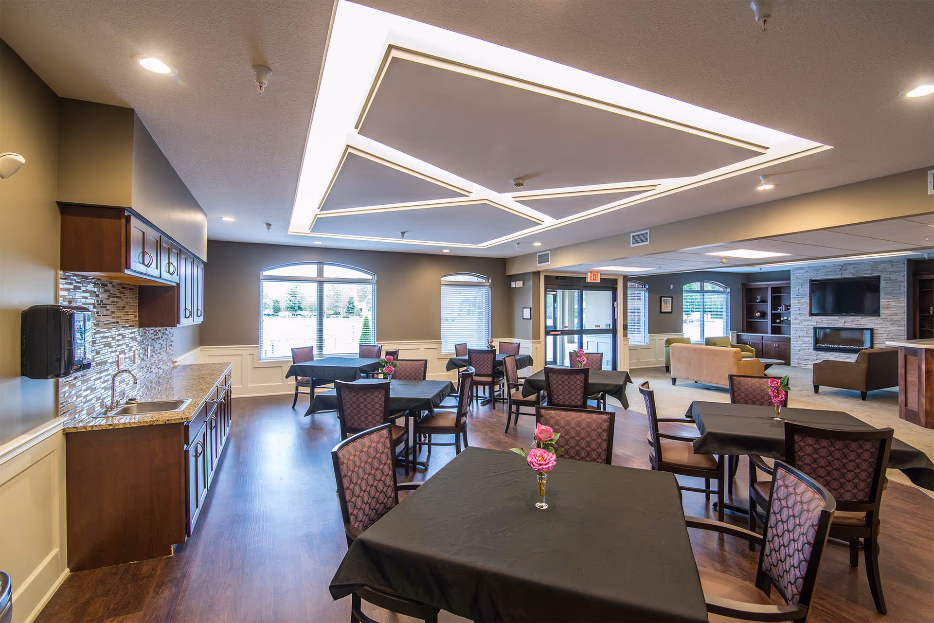 Spacious senior living dining and lounge area with multiple tables and chairs, a kitchenette on the left, and seating with a TV and fireplace in the background.