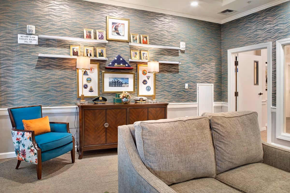 A cozy living room area with a gray couch in the foreground and a blue armchair with floral sides and an orange pillow to the left. Against the wall is a wooden cabinet topped with two lamps and various framed photos and memorabilia, including a folded American flag in a triangular display case. The wall behind has a wavy patterned wallpaper, and there are two open doorways on the right side of the image.