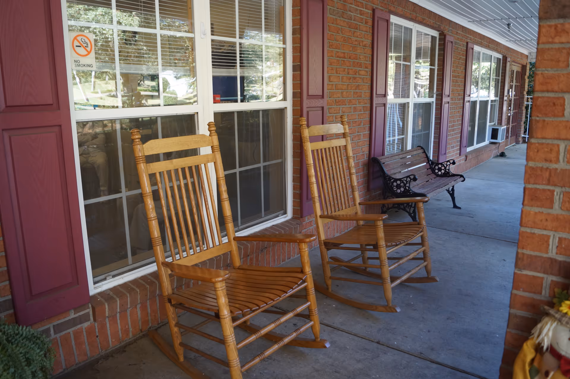 A covered outdoor porch area with two wooden rocking chairs and a metal bench with wooden slats. The porch has a brick wall with windows featuring purple shutters. A 'No Smoking' sign is visible on one of the windows.