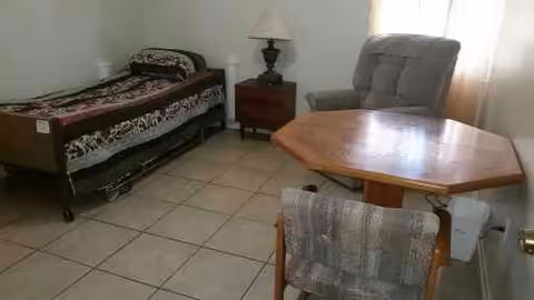 Small bedroom with a single bed, bedside table and lamp, recliner, and a wooden table with a chair on a tiled floor.