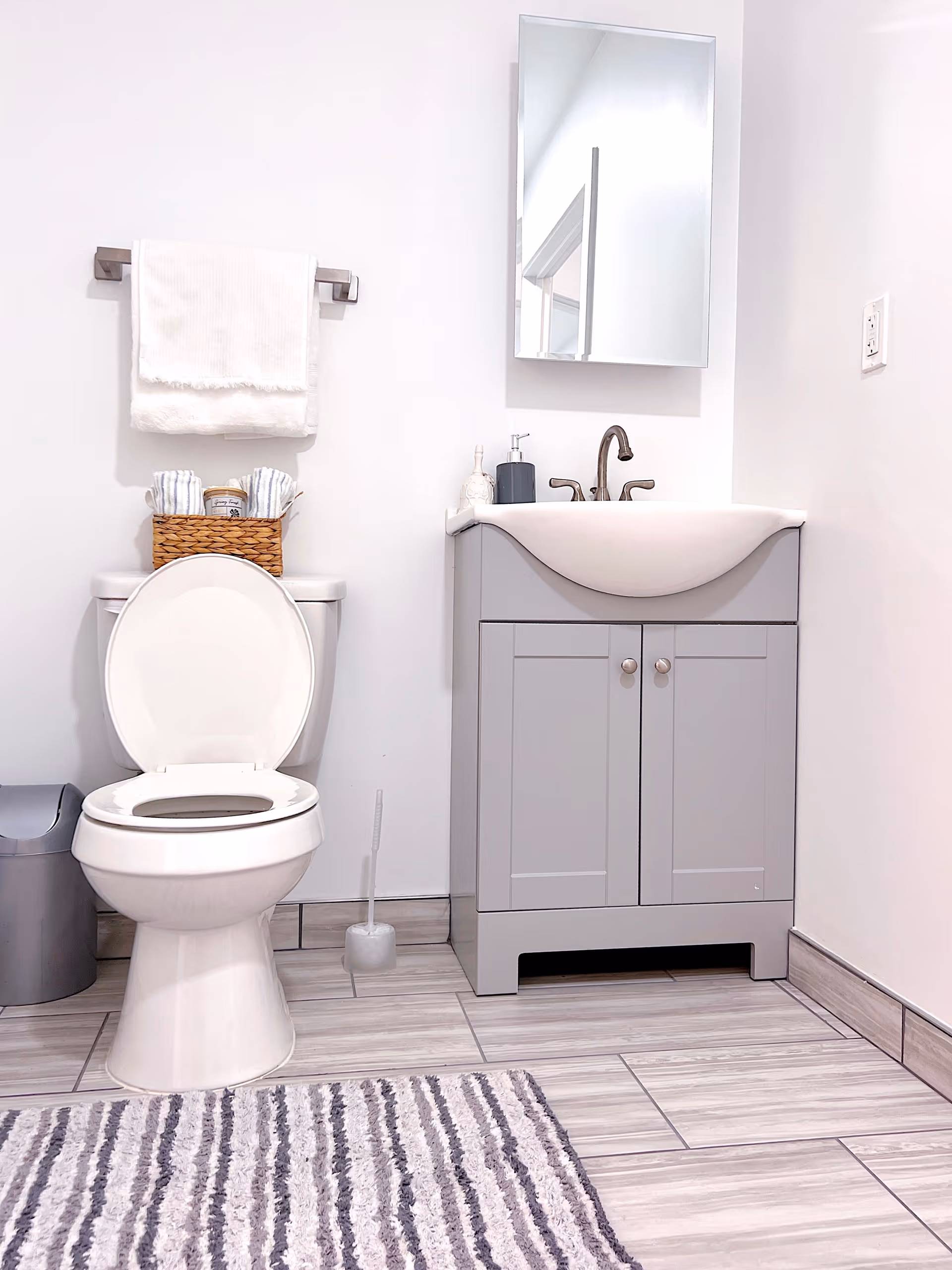 A clean and modern bathroom featuring a white toilet with the lid open, a gray vanity with a white sink and bronze faucet, a rectangular mirror above the sink, a towel rack with a white towel, a small wicker basket with rolled towels and a candle on top of the toilet tank, a gray trash can, a toilet brush, and a striped gray and white rug on the tiled floor.
