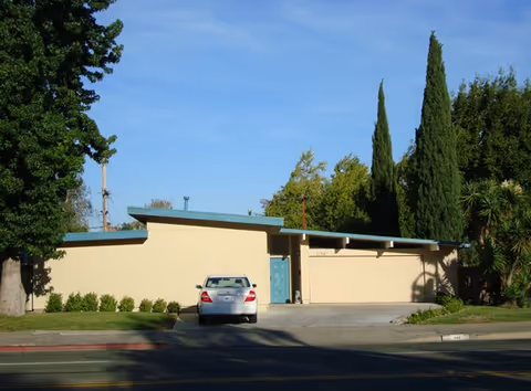 Single-story building with a flat roof and a blue front door, surrounded by trees and bushes, with a silver car parked in the driveway in front of the building.