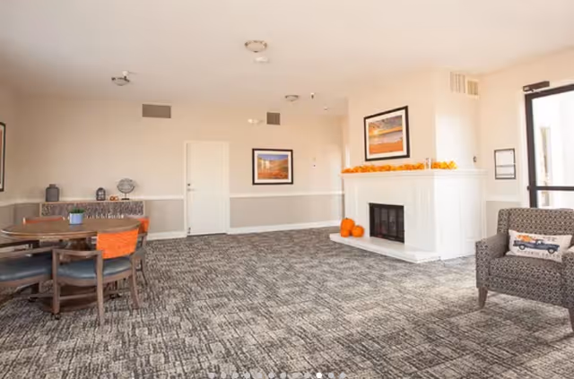 A spacious senior living common area with patterned carpet flooring, a white fireplace decorated with small pumpkins and orange garland, a framed landscape painting above the fireplace, a round wooden table with chairs on the left side, and a patterned armchair with a decorative pillow on the right side near a glass door.