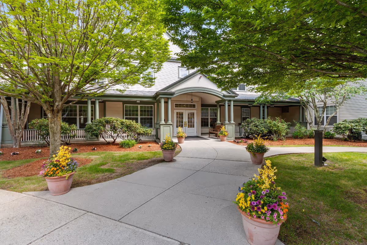 Entrance to a senior living facility with a covered porch supported by green columns, surrounded by well-maintained landscaping including trees, bushes, and flower pots with colorful flowers along a curved concrete walkway.