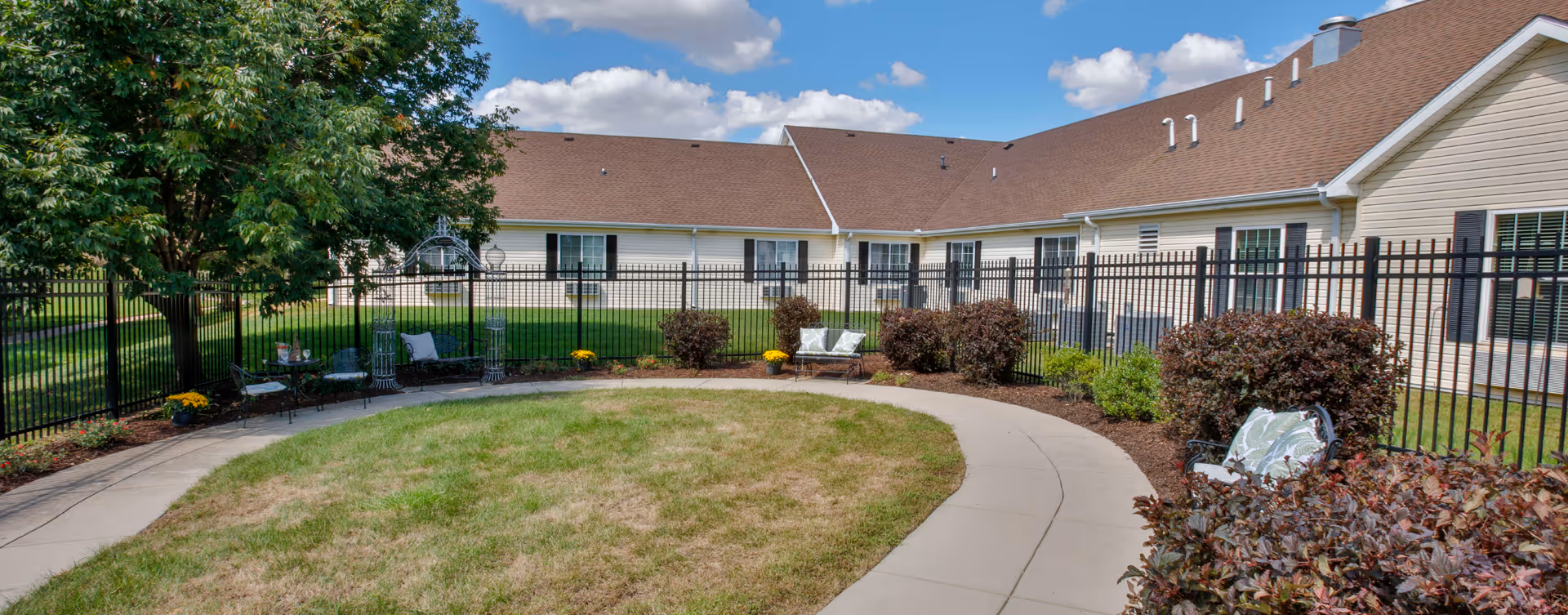Outdoor courtyard area at Bickford of Champaign featuring a curved concrete walkway surrounding a grassy lawn, black metal fencing, several benches with cushions, bushes, a large tree, and a beige building with multiple windows under a partly cloudy blue sky.