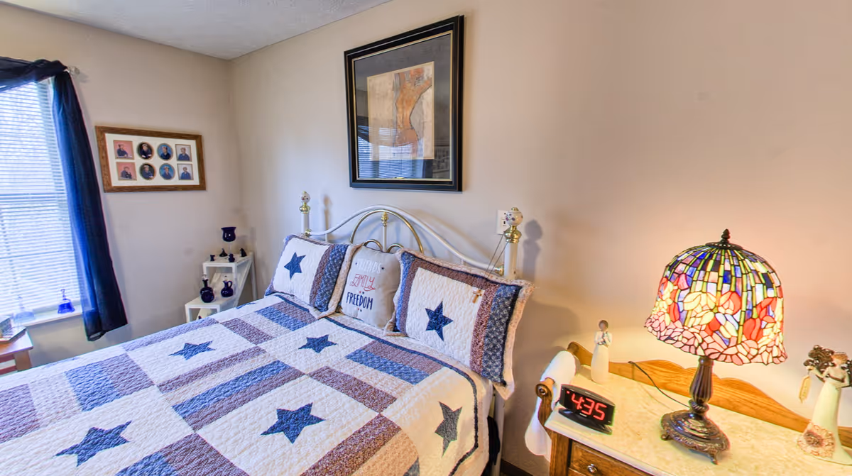 Small furnished bedroom with a quilted star-pattern bed, framed art on the wall, a bedside table with a stained-glass lamp and digital clock, and a curtained window.