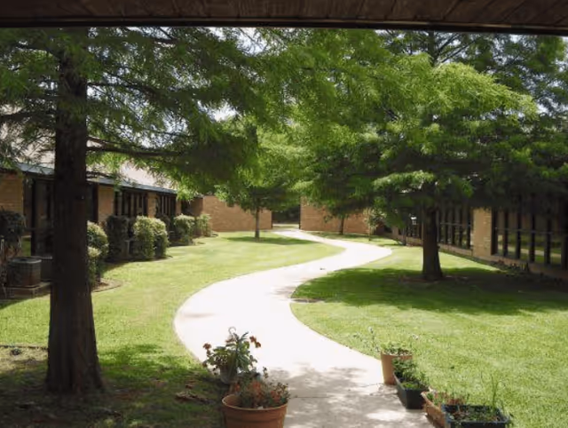 A winding concrete pathway through a green lawn with several trees and potted plants, bordered by single-story brick buildings with windows on either side.